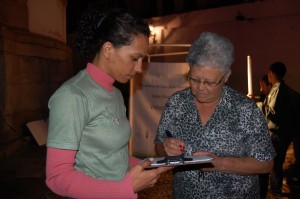 03dia-de-ns-rosa_rio-op A depoente Maria Aparecida Santos, assina o Termo de Autorização para uso de imagem, ao lado de Cristiane Evangelista, no adro da Igreja Nossa Senhora do Rosário dos Pretos, no bairro Rosário, em Ouro Preto. Foto: Marianne Fonseca
